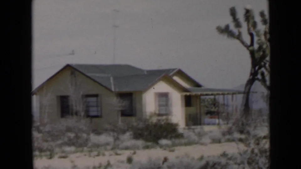 San Diego-area 1949 frame of a modest single-story house with a covered porch, dry brush, and a tree-like desert plant at right.