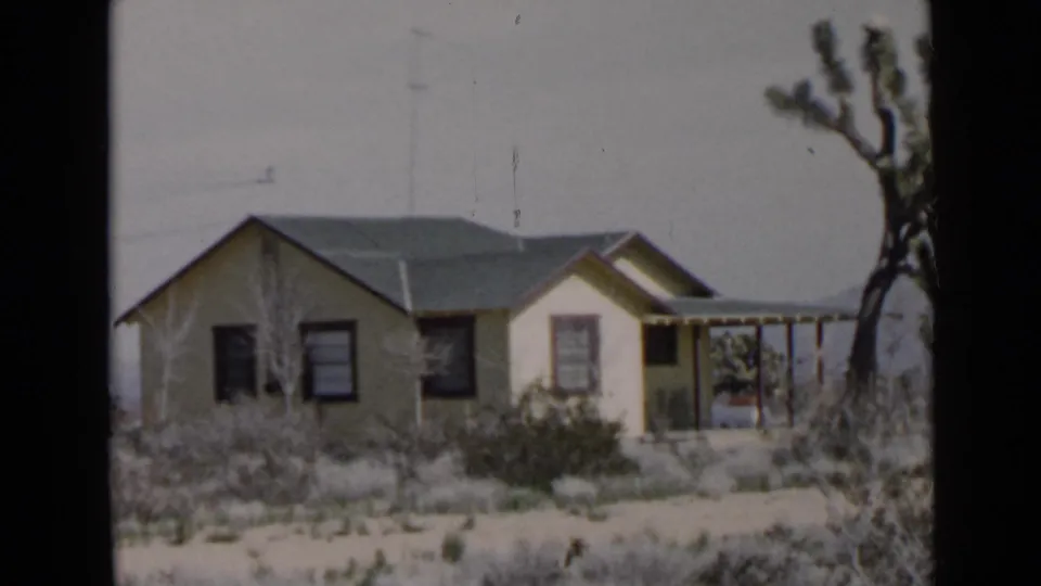 San Diego-area 1949 home-movie frame centering the small house, porch, dark windows, dry vegetation, and a Joshua tree-like plant.