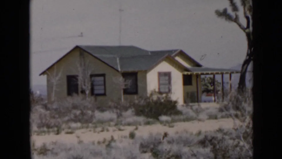 San Diego-area 1949 frame showing the small house from a slightly wider angle, with porch, dry yard, and desert plant at the right edge.