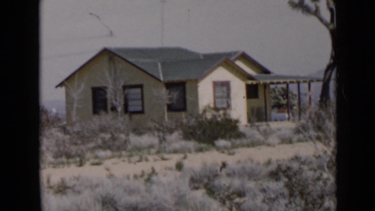 Small yellow house in dry scrubland with a dark roof and hazy hills beyond.