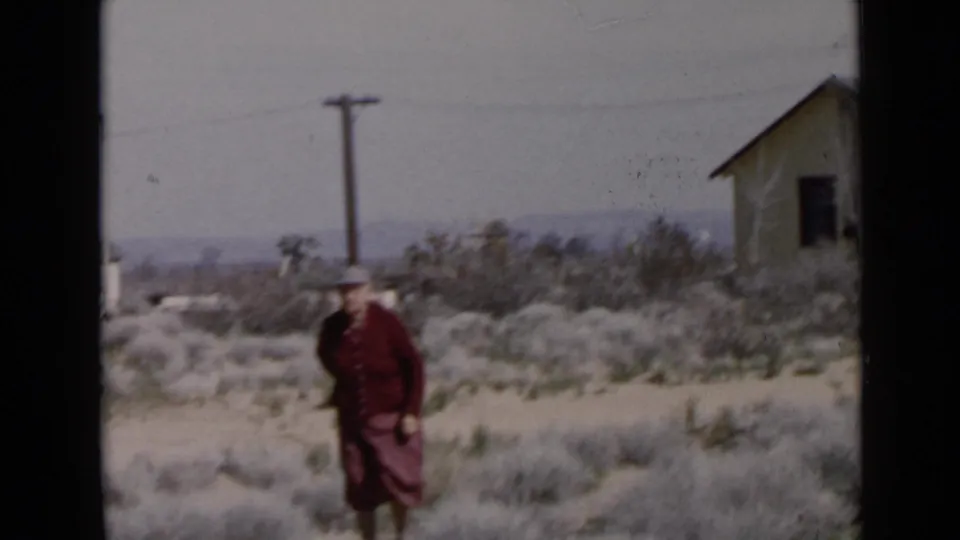 1949 San Diego-area home-movie frame showing an adult woman in a red suit walking through dry scrub near a small house.