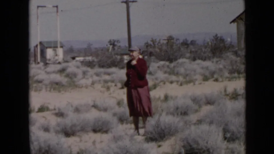 1949 San Diego-area frame of an adult woman in a red suit and hat in dry terrain, with utility poles and a house nearby.