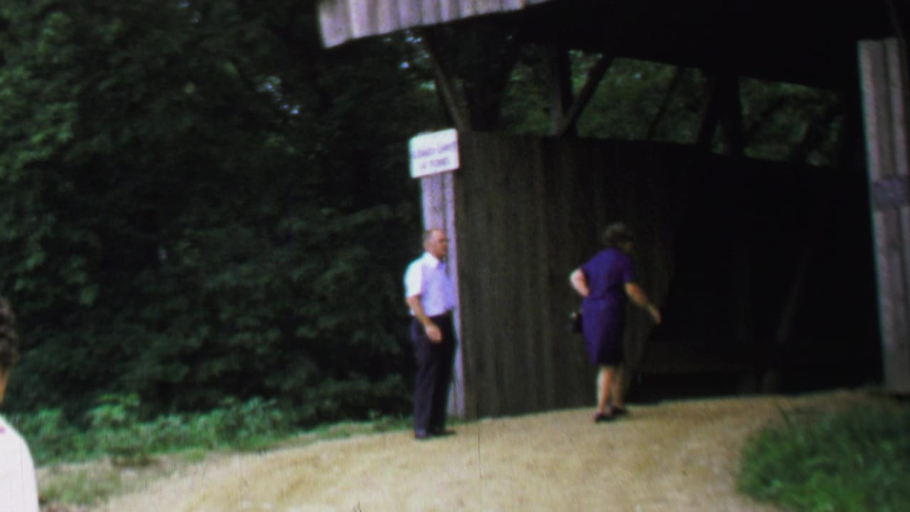 1969 Washington DC home-movie frame showing a woman approaching a covered bridge entrance as a man waits near posted signs.