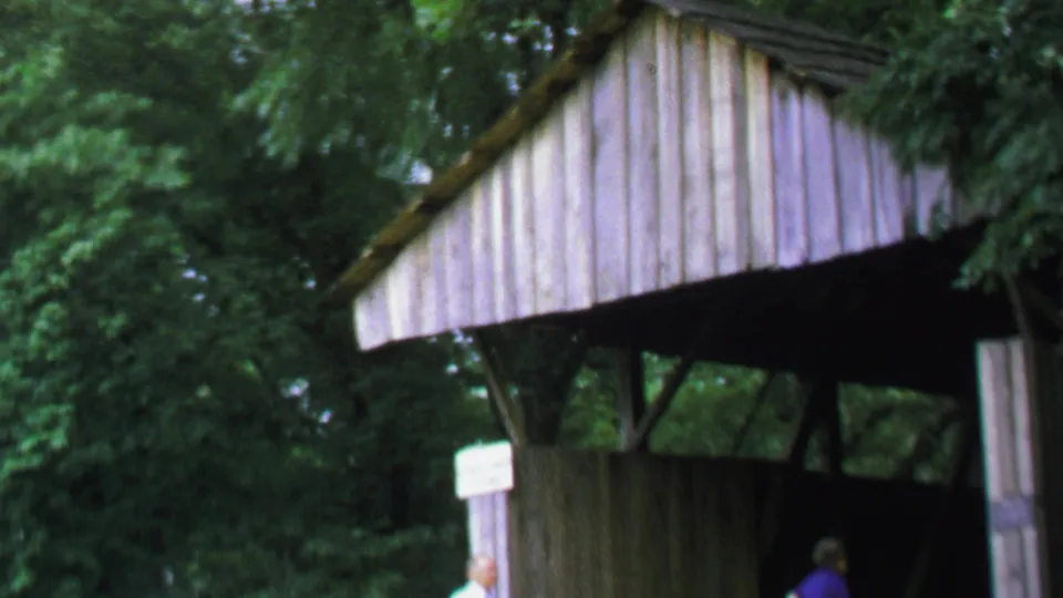 The camera shifts upward on the rustic covered bridge entrance, with trees around it and the man partly cropped below.