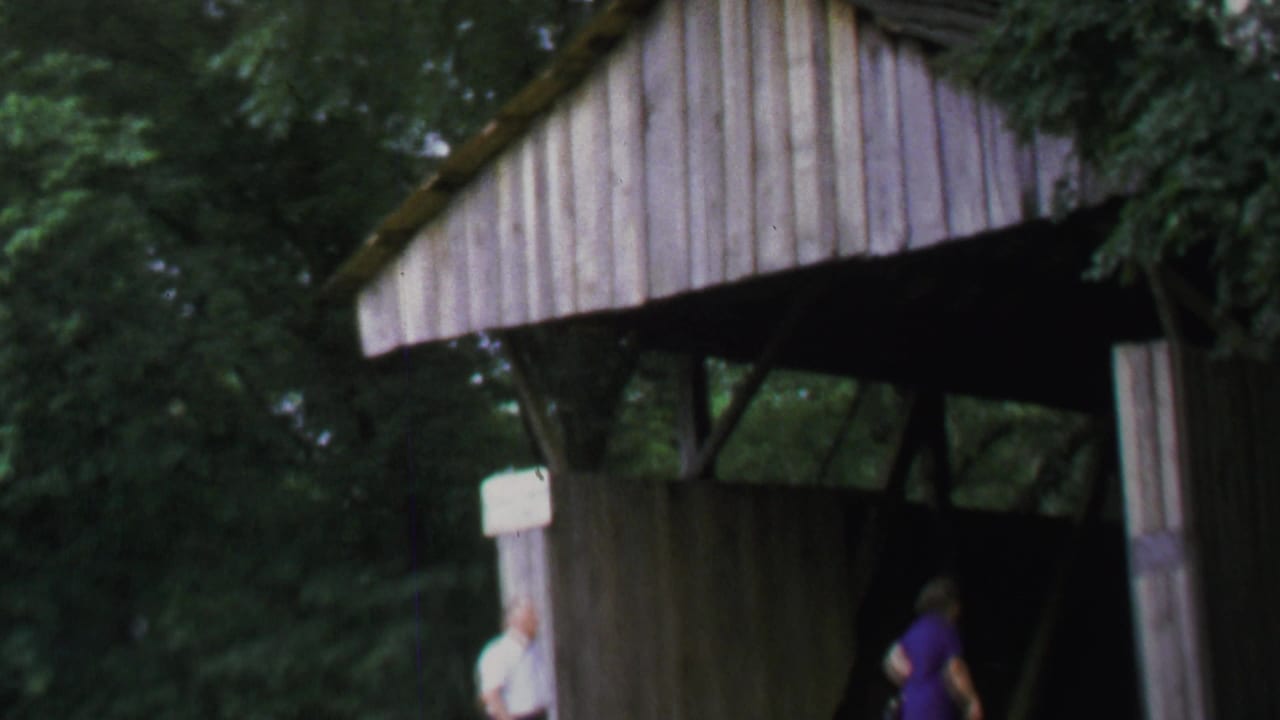1969 home-movie frame showing adults approaching a wooden covered bridge surrounded by trees in Washington, DC metadata context.