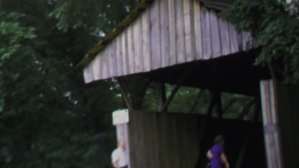 1969 home-movie frame showing adults approaching a wooden covered bridge surrounded by trees in Washington, DC metadata context.