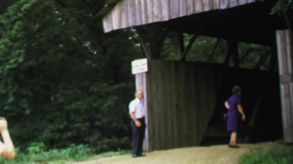 A man in a light shirt stands by the covered bridge entrance while a woman in purple walks into the shaded interior.