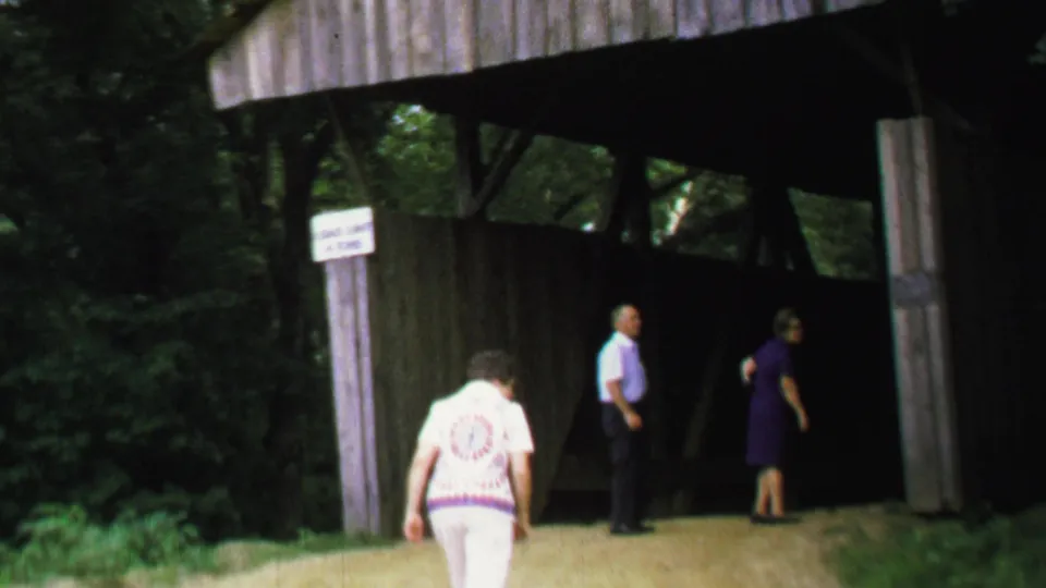 Three adult visitors are visible near a wooden covered bridge, with one person in white walking toward the entrance.