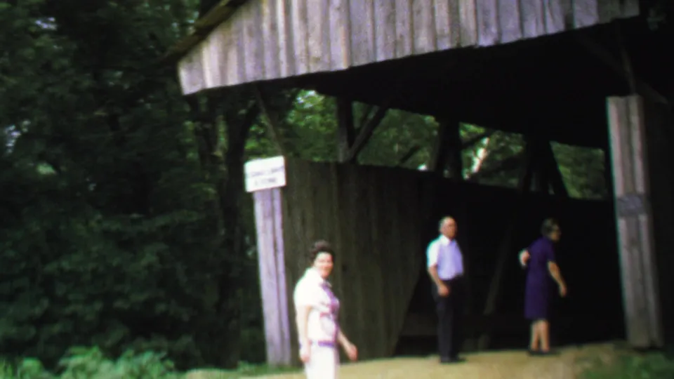 A woman in a light patterned outfit looks toward the camera while two adults remain near the covered bridge entrance.
