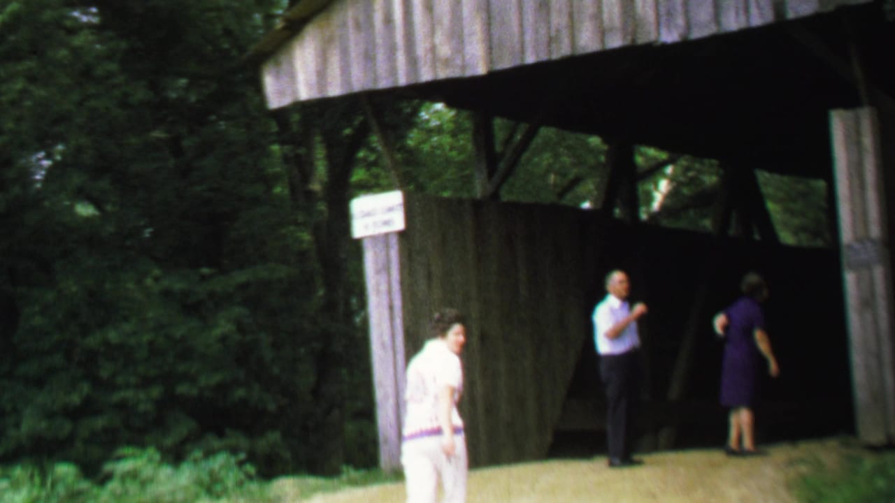 1969 Washington, DC home-movie frame showing adults near a rustic wooden covered bridge, with a sign on the entrance post.