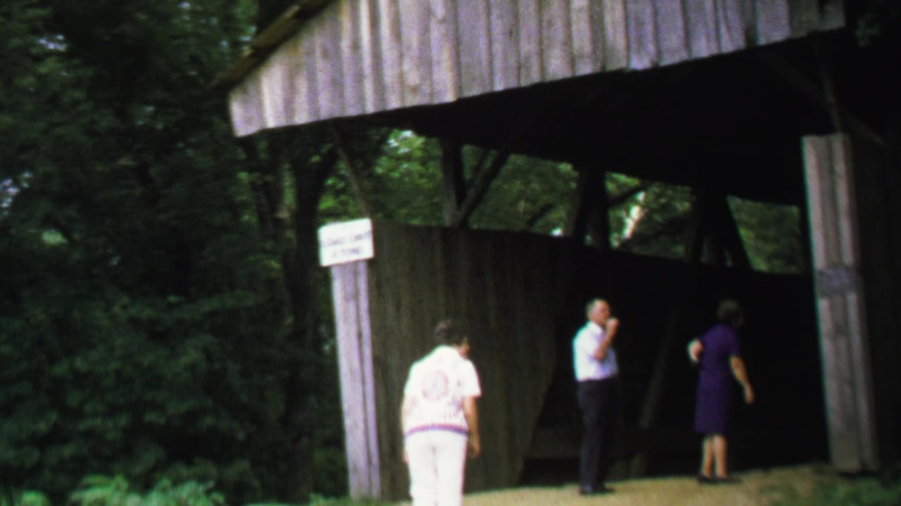 Adults stand and walk at a covered bridge entrance beside a wooded area.
