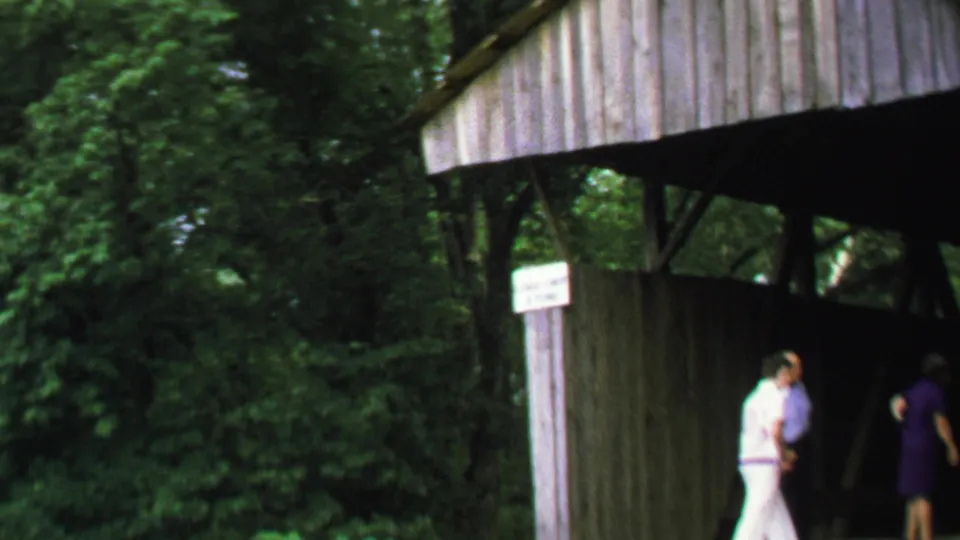 1969 Washington, DC home-movie frame showing visitors moving around the entrance of a wooden covered bridge in trees.