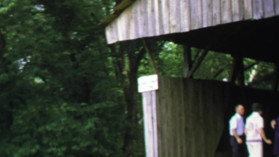 1969 Washington, DC frame with adults partly at the edge of view beside a rustic covered bridge and surrounding trees.