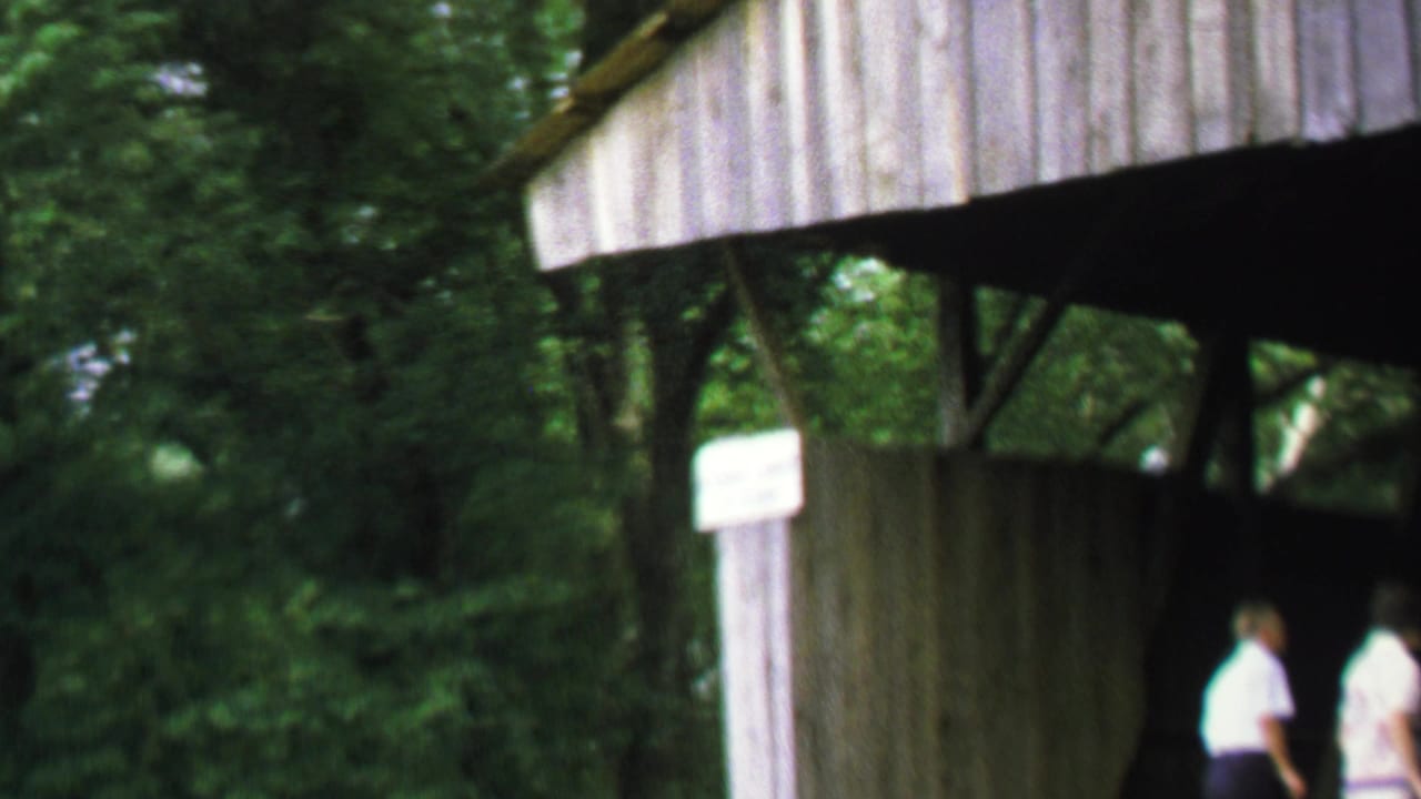 1969 home-movie frame of adults near a wooden covered bridge entrance in a leafy Washington, DC metadata context.