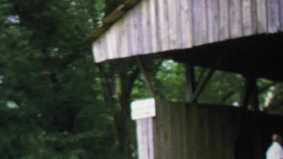 Soft 8mm frame showing a person by the shaded opening of a wooden covered bridge, with trees around the structure.