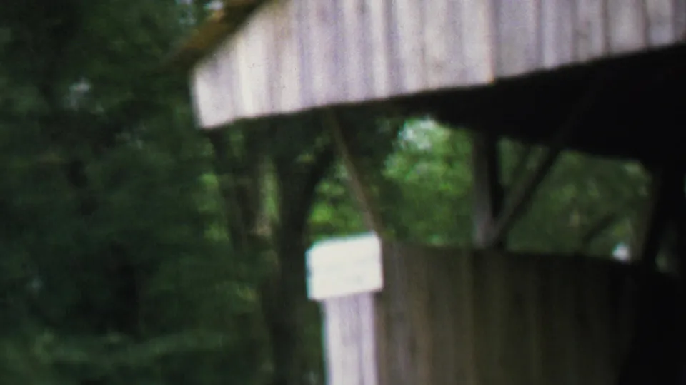 Blurred 8mm close view of the covered bridge exterior, with green trees and a small sign beside the entrance.