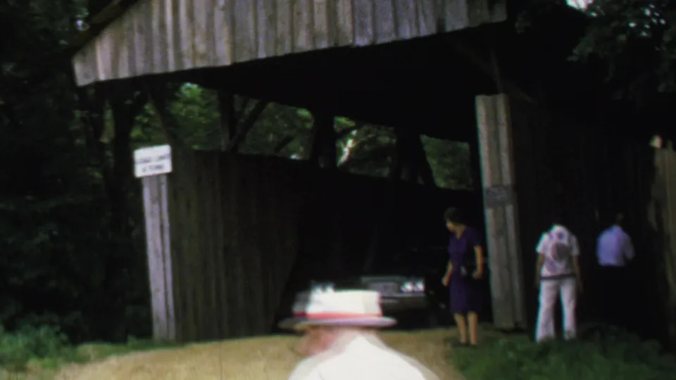 1969 8mm frame with adults and a light-colored car inside a shaded wooden covered bridge.