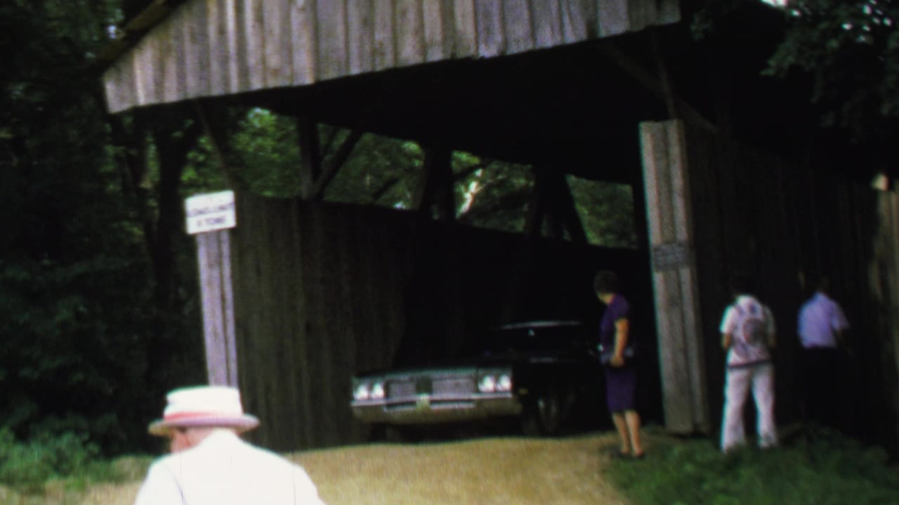 1969 8mm home-movie frame showing a dark American sedan partly inside a wooden covered bridge, with adults standing nearby in a wooded setting.