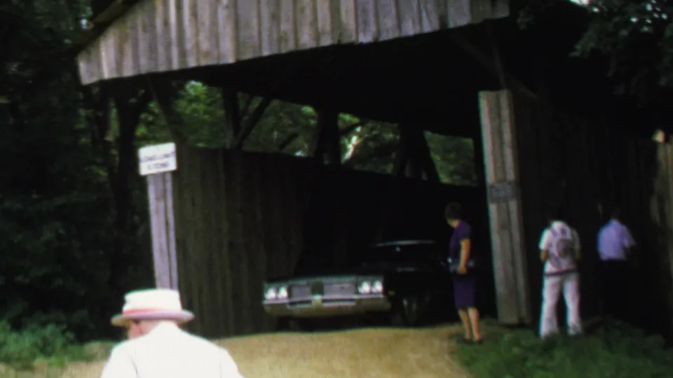 1969 8mm home-movie frame showing a dark American sedan partly inside a wooden covered bridge, with adults standing nearby in a wooded setting.