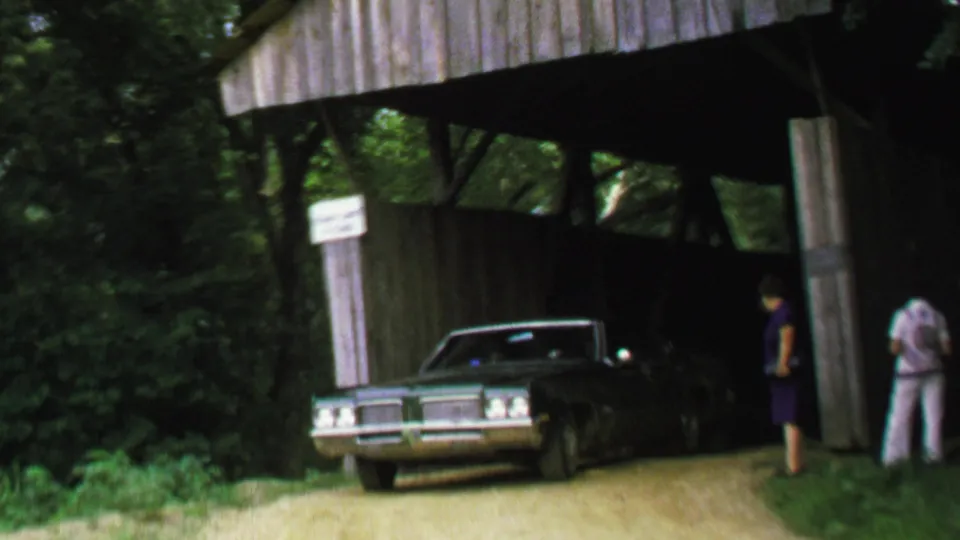A dark late-1960s style sedan comes out of the covered bridge onto the dirt road as adults stand near the wooden wall and foliage.