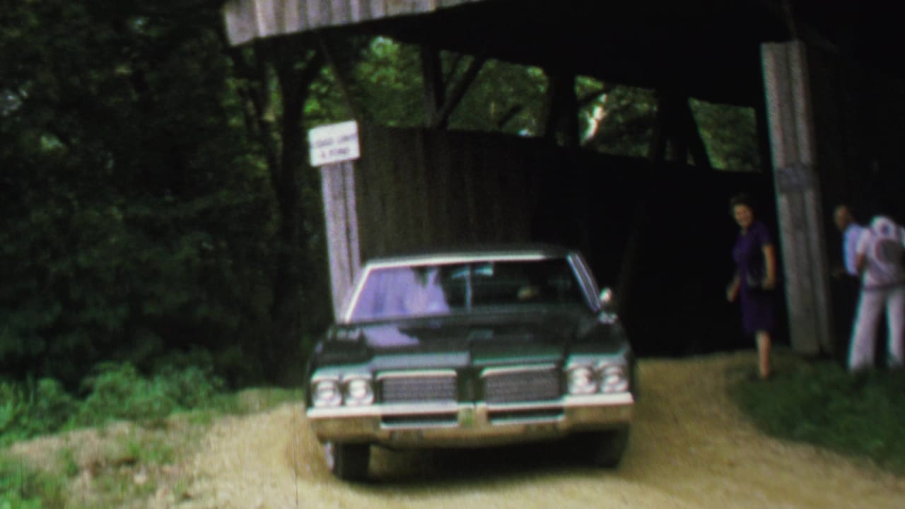 1969 8mm frame of a green sedan nearing a covered bridge entrance with several adults beside the road.