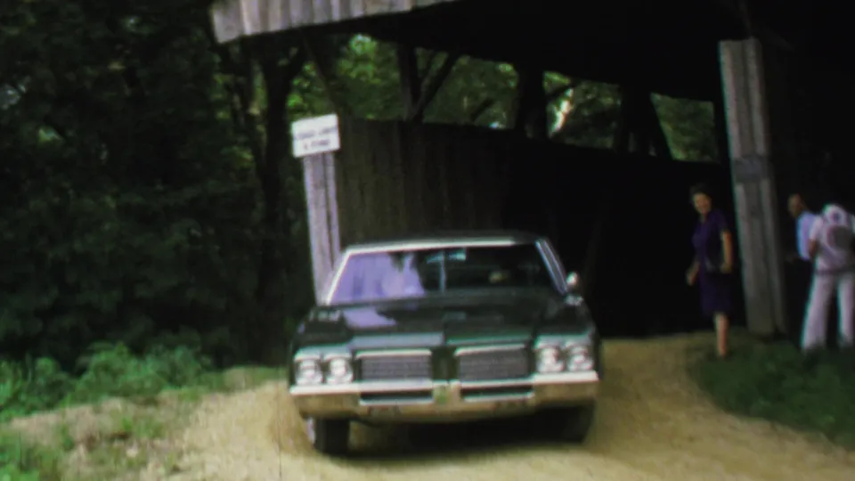 1969 8mm frame of a green sedan nearing a covered bridge entrance with several adults beside the road.