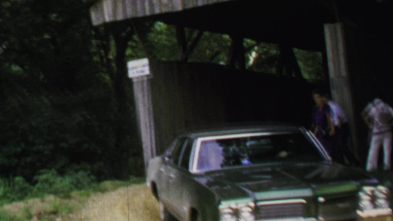 The green sedan moves partly out of frame near the covered bridge, with adults visible by the entrance and roadside vegetation.