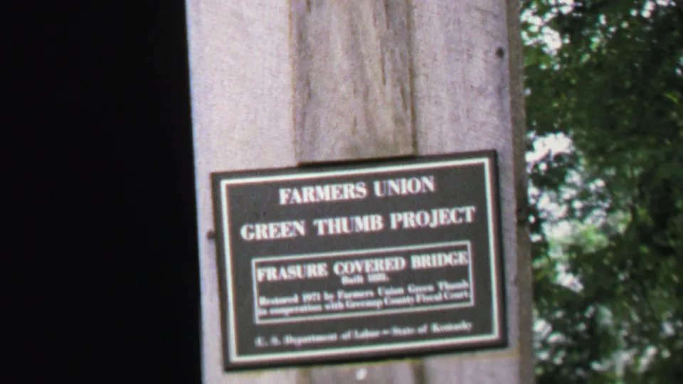 Close view of a bridge plaque reading Farmers Union Green Thumb Project and Frasure Covered Bridge on a wooden post.