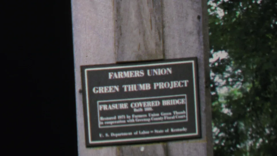 The plaque for Farmers Union Green Thumb Project and Frasure Covered Bridge is shown against the wooden bridge post and trees.