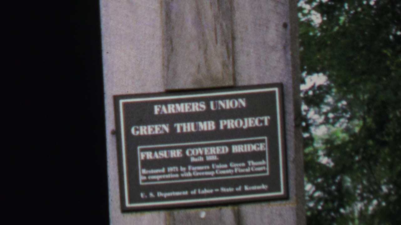 Close view of a Farmers Union Green Thumb Project sign for Frasure Covered Bridge on a wooden post, with trees behind it.