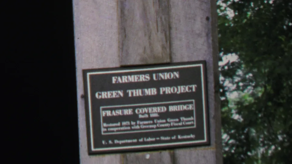 Close view of a Farmers Union Green Thumb Project sign for Frasure Covered Bridge on a wooden post, with trees behind it.