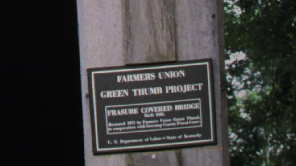The Frasure Covered Bridge project sign remains centered on a timber support, with dark bridge shadow and leafy background visible.
