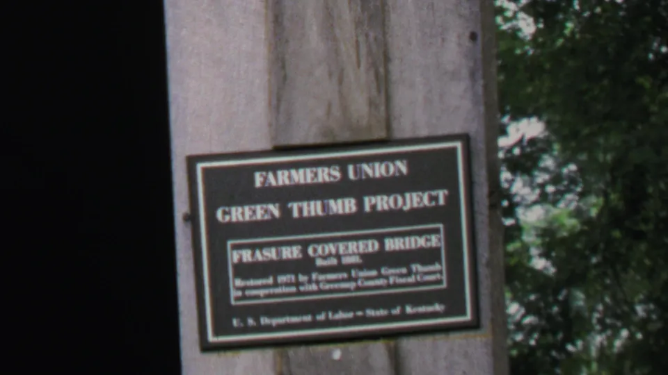 Handheld 8mm view of the Farmers Union Green Thumb Project sign, with the Frasure Covered Bridge text legible in the center panel.