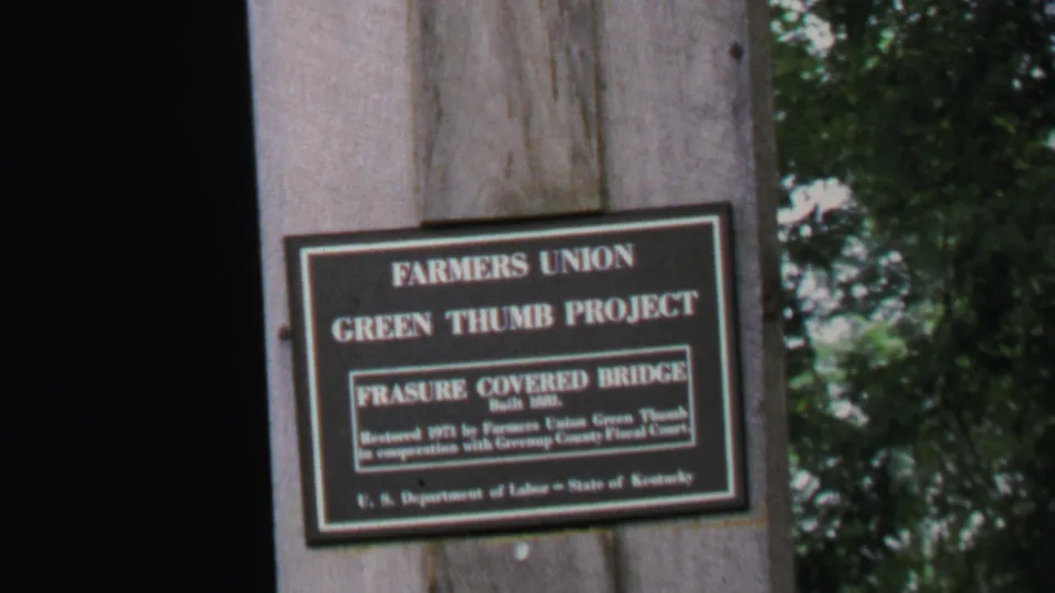 Final frame holds on the Green Thumb Project marker for Frasure Covered Bridge, mounted to wood with foliage at the edge of frame.