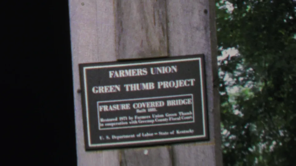 1969 home-movie frame showing a Farmers Union Green Thumb Project sign near a wooded road with people and parked cars in the distance.