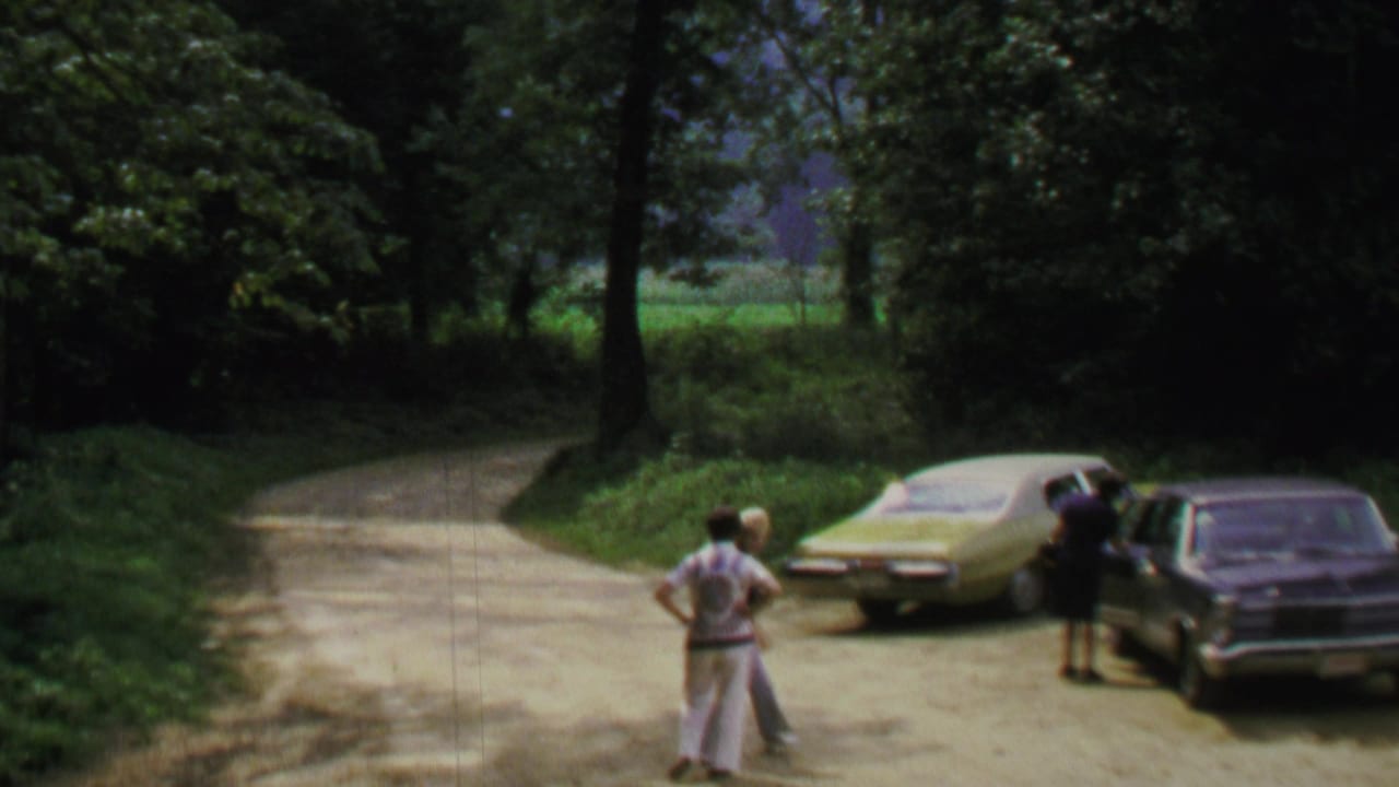 Three adults stand near two parked sedans on a shaded dirt road.
