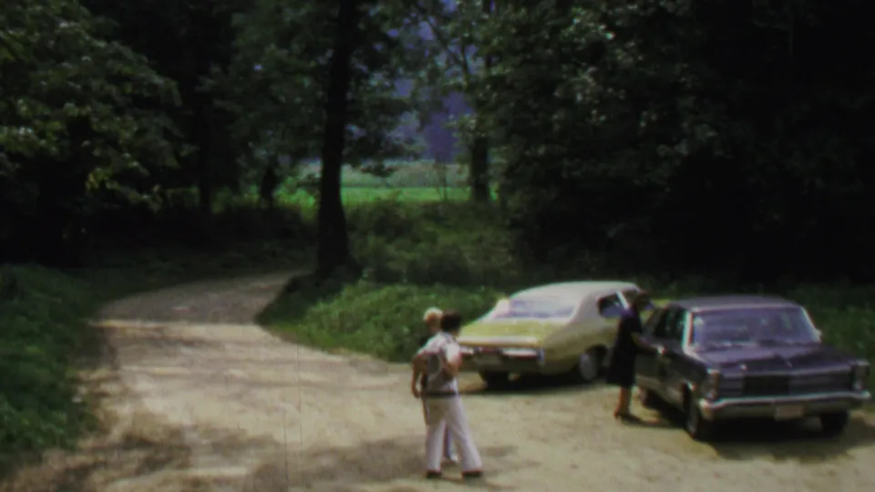 1969 Washington DC home-movie frame with adults standing apart near a yellow car and dark sedan beside a wooded road.