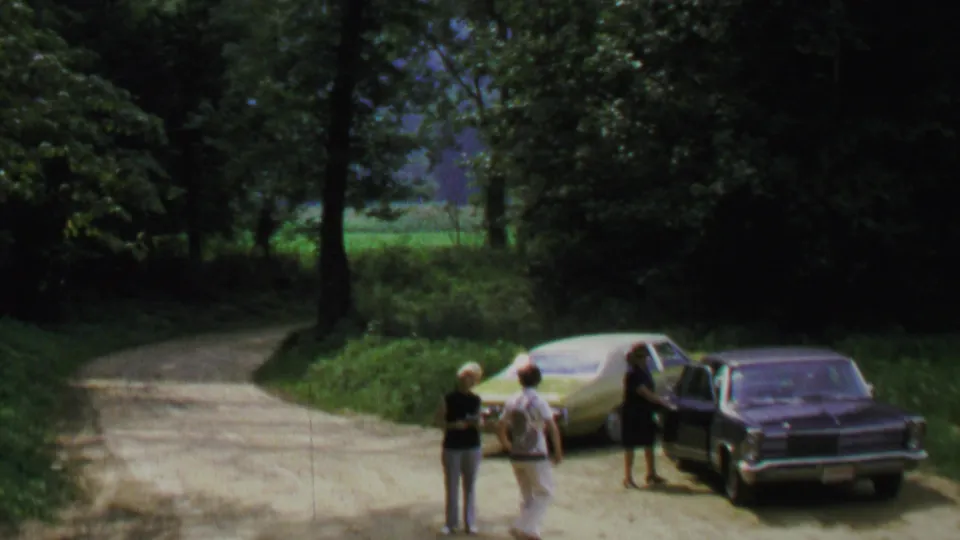 1969 Washington DC home-movie frame showing three adults beside parked sedans at a shaded bend in a wooded dirt road.