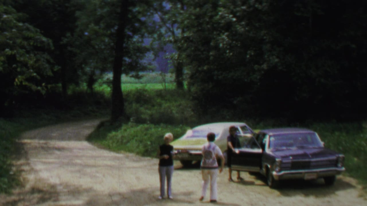 1969 home-movie frame shows adults near light and dark sedans on a wooded dirt road in the Washington, DC metadata location.