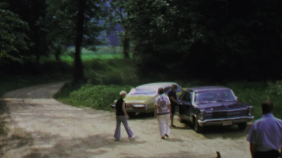 1969 home-movie view of adults moving around two parked sedans on a rural road, with another figure partly visible in the foreground.
