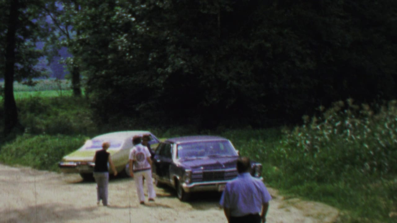 1969 8mm home-movie frame near Washington, DC showing adults beside two parked sedans on a wooded dirt road.