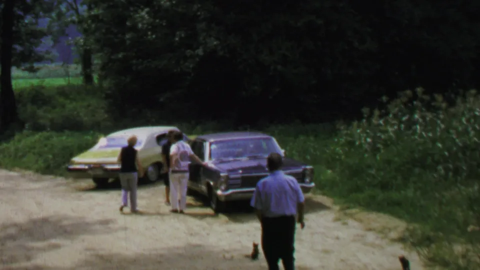 1969 Washington, DC area home-movie frame with adults gathered beside light and dark sedans in a wooded roadside clearing.