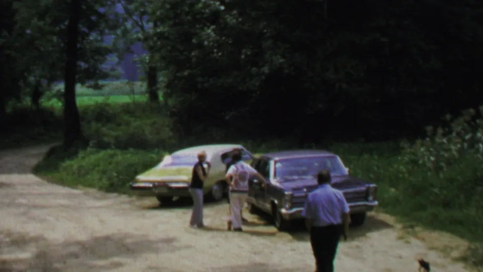 Late-1960s 8mm frame showing adults talking between parked sedans on a rural-looking road in the Washington, DC area.