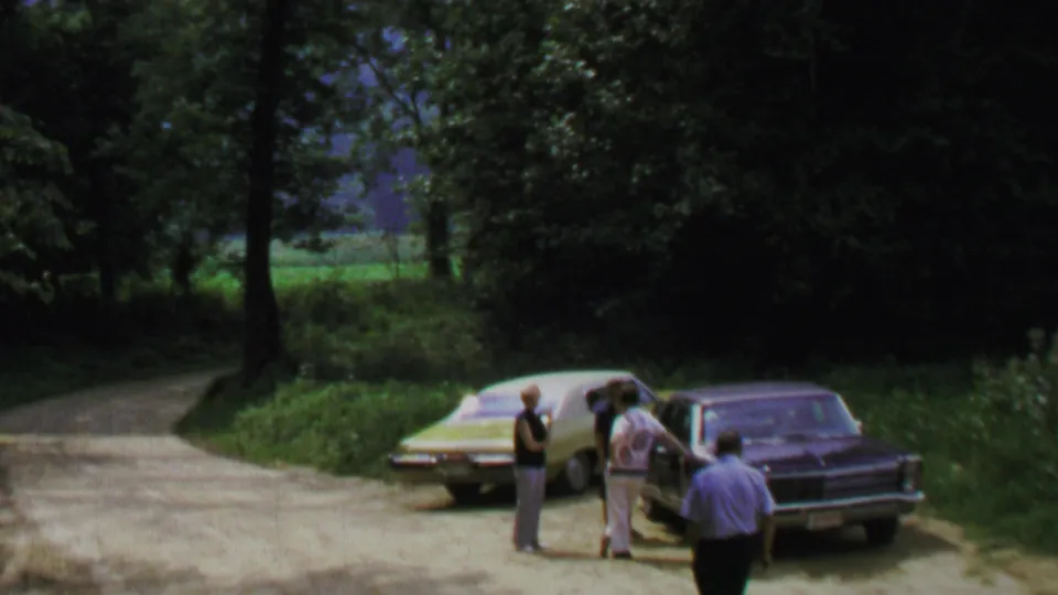 1969 home-movie frame near Washington, DC with a small group beside parked sedans and a wooded roadside behind them.