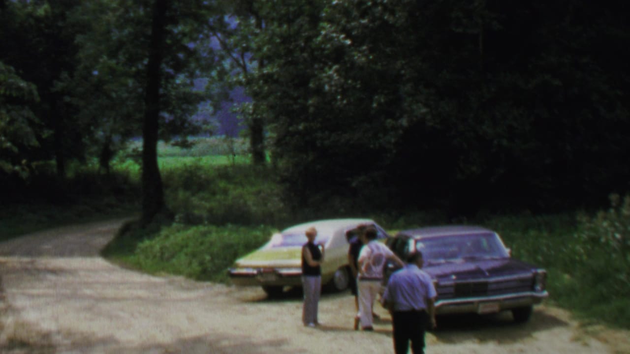 1969 Washington, DC-area home-movie frame showing adult men beside parked cars on a shaded roadside with trees and grass.