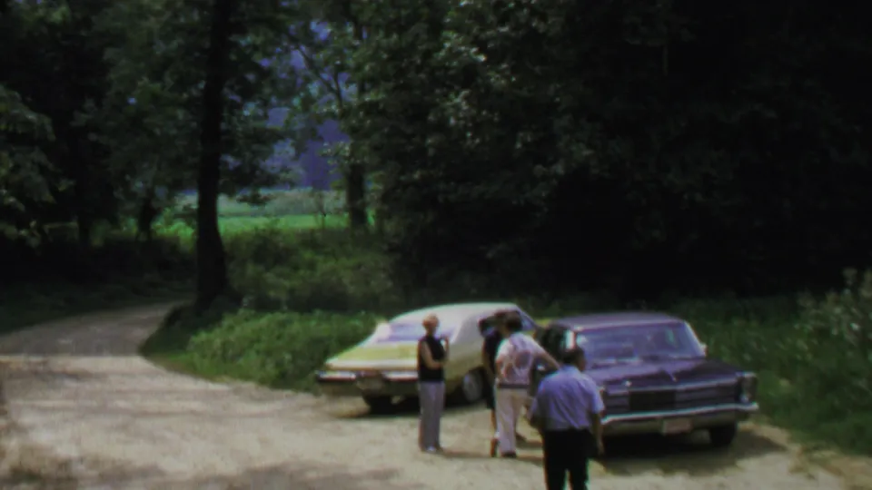 1969 Washington, DC-area home-movie frame showing adult men beside parked cars on a shaded roadside with trees and grass.