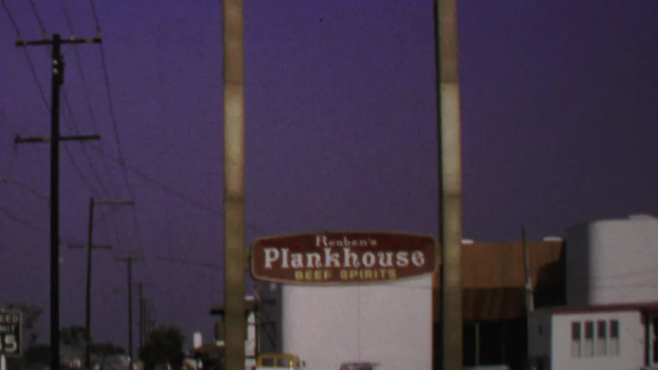 1969 Washington, DC 8mm frame showing the Reuben's Plankhouse sign, cars near the building, roadside signs, utility poles, and wires.