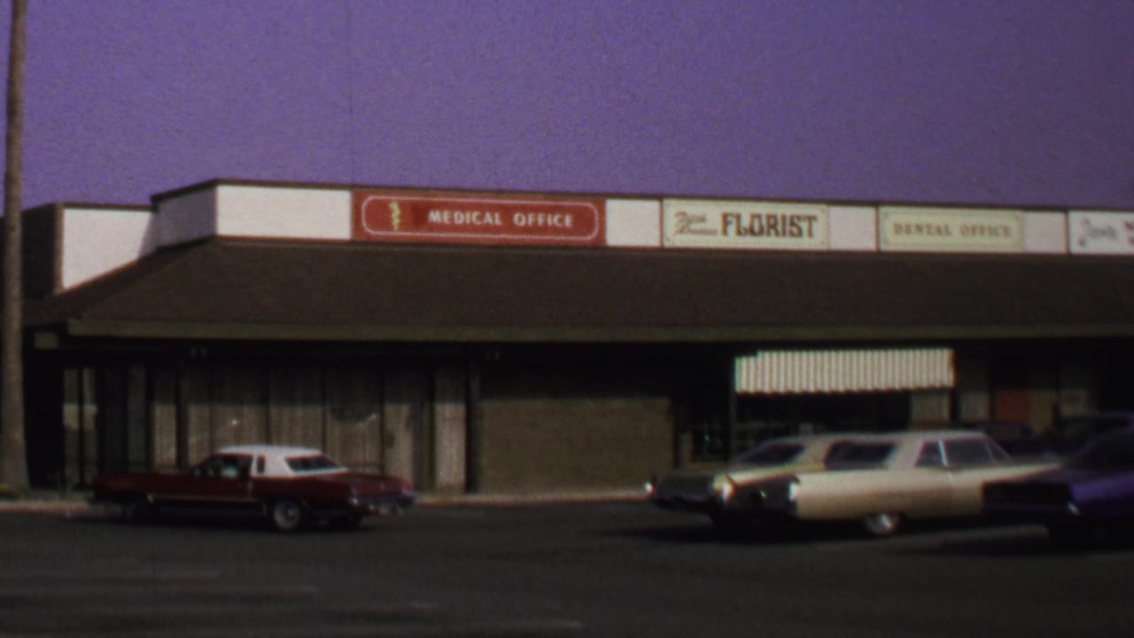 1969 Washington, DC home-movie frame showing a red two-door car near storefronts marked medical office, florist, and dental office.