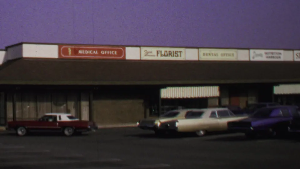 1969 Washington, DC home-movie frame showing the red two-door car reversing near a cream sedan outside a mid-century commercial strip.