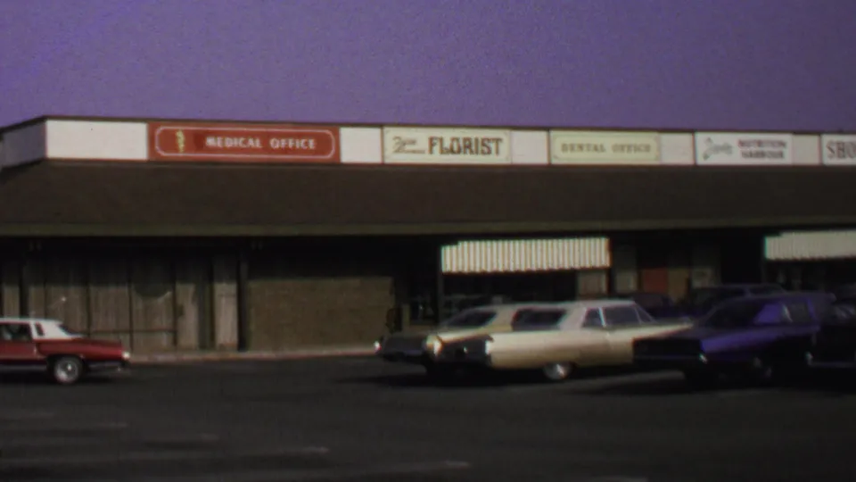 1969 Washington, DC frame with a cream sedan and dark car in front of a strip center showing medical office, florist, and dental signage.